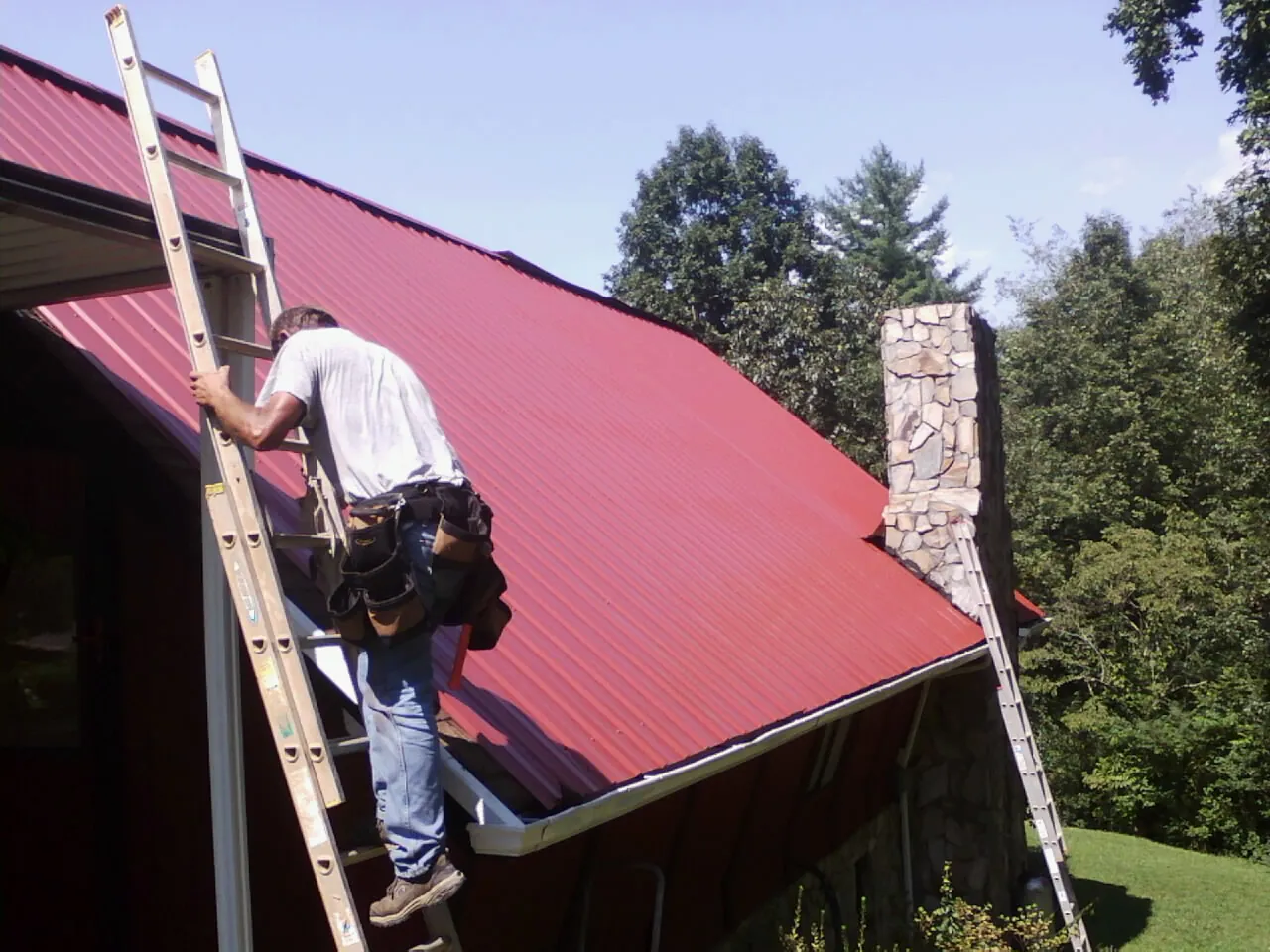 Roofer on a ladder beside a red metal roof and stone chimney — Soffit & Fascia Repair by Roofing Brothers in Oakmont